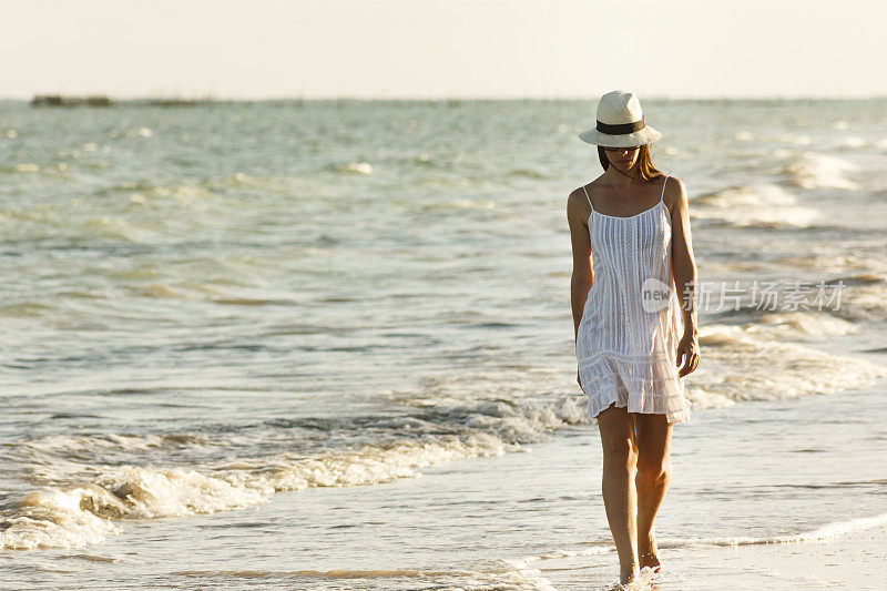 Portrait of beautiful woman walking at the beach at sunset in Alagoas, Northeastern Brazil. She is wearing white dress, sunglasses and straw hat